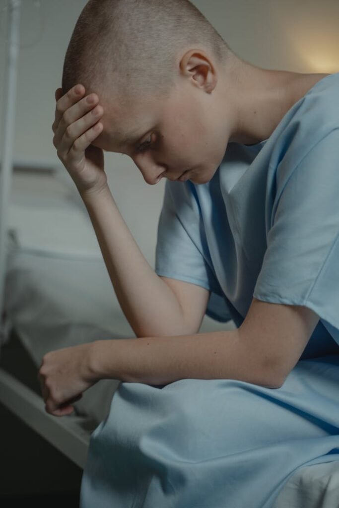 Bald woman in a hospital gown, sitting and looking down, symbolizes illness and contemplation.