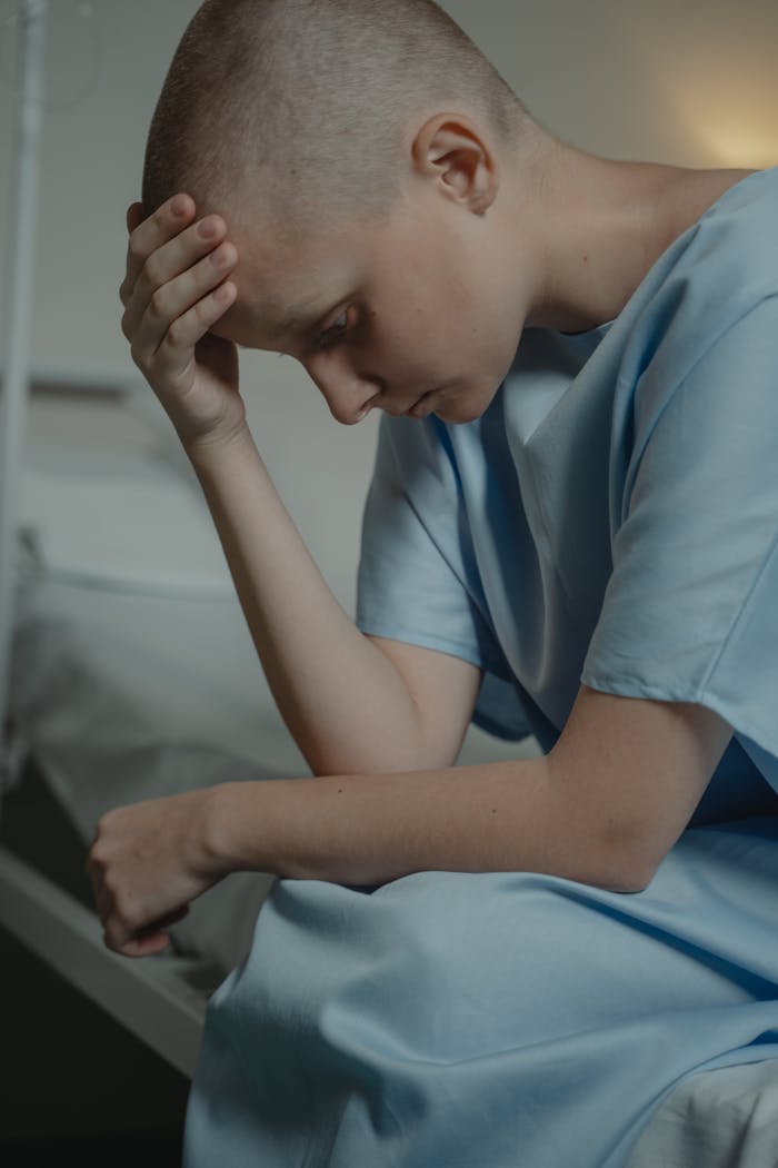 Bald woman in a hospital gown, sitting and looking down, symbolizes illness and contemplation.